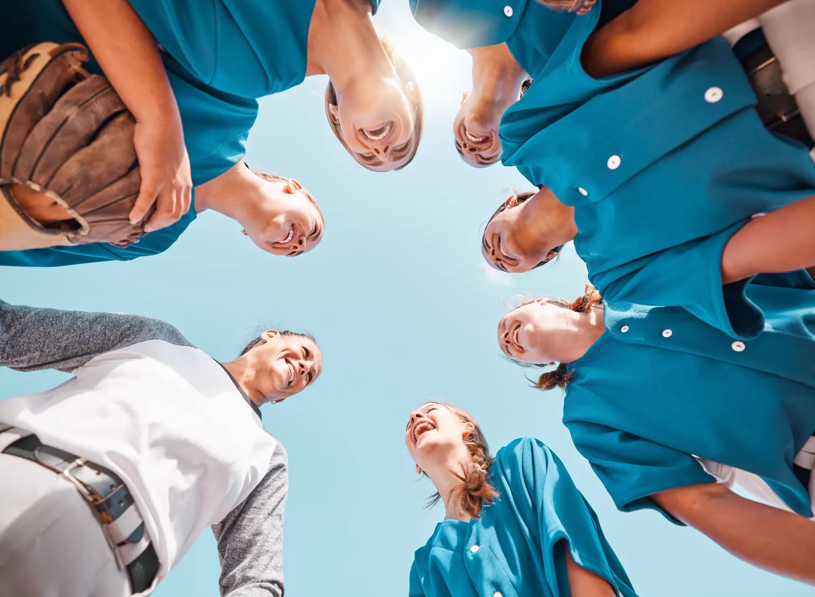 Young softball players in a team huddle looking down and smiling under a bright sky