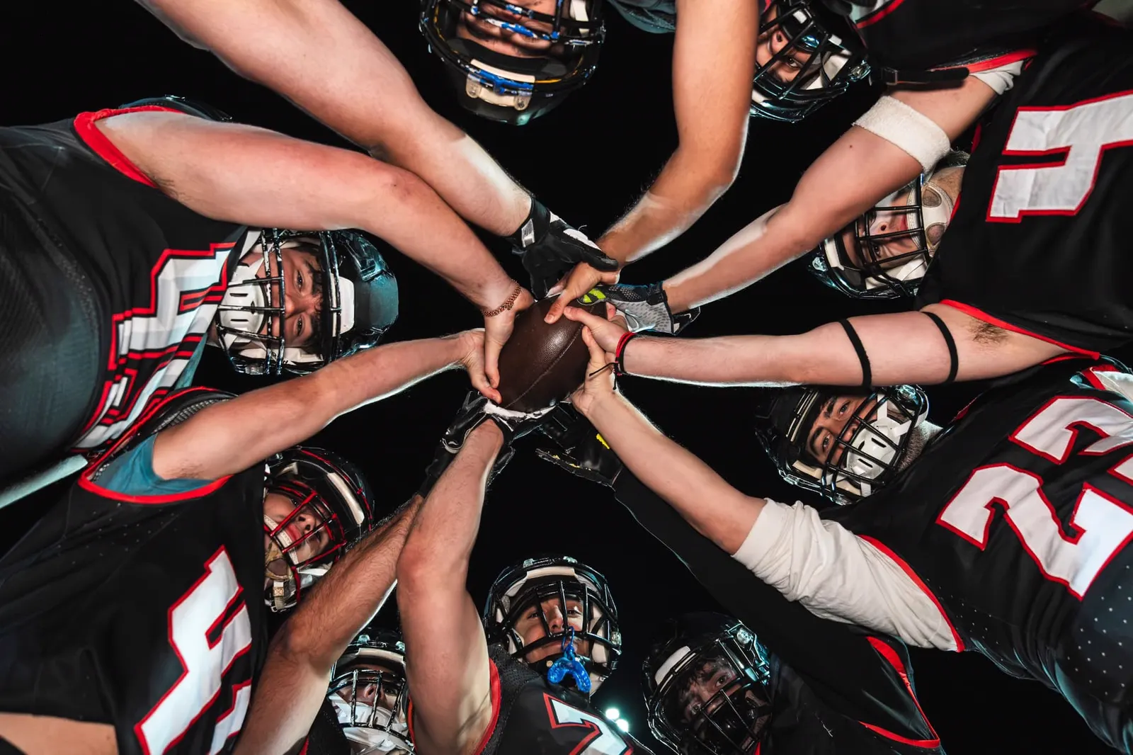 Football players in a huddle reaching their hands together over a football against a night sky