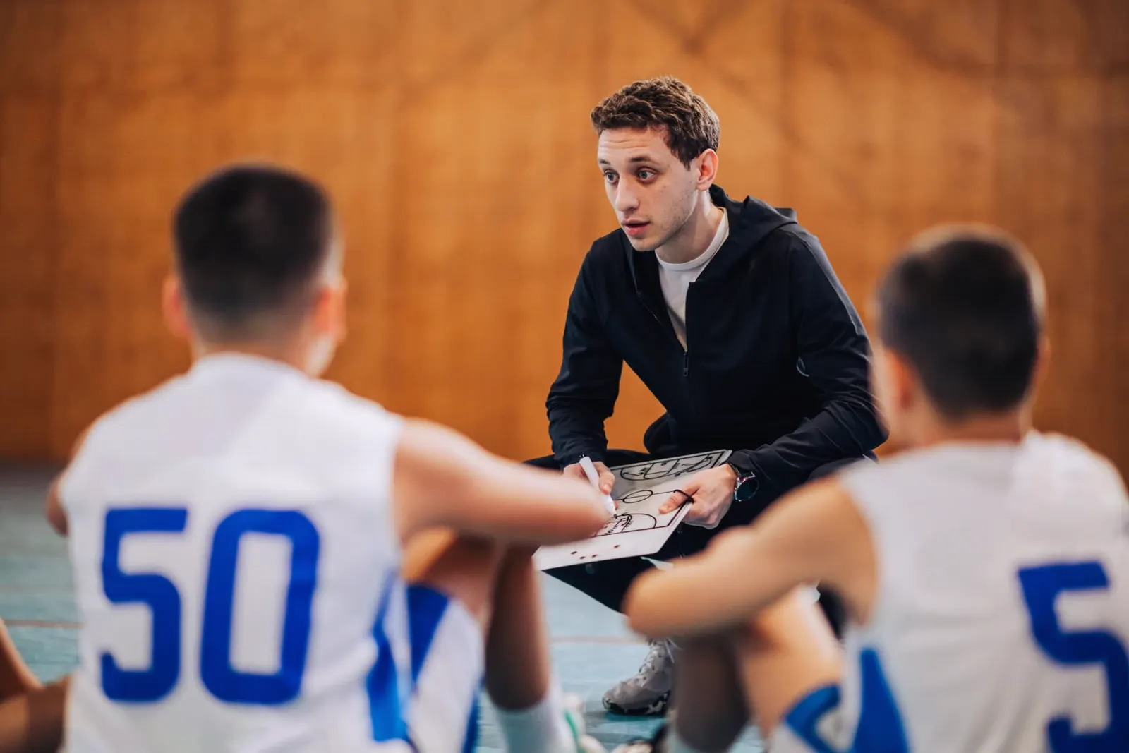 Basketball coach crouching to explain plays on a whiteboard to two young players sitting on the court